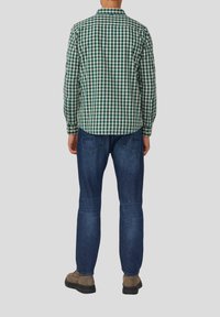 Man in een groen-wit geruit overhemd, blauwe jeans en bruin schoenen, staand met zijn gezicht naar weg tegen een egale achtergrond.