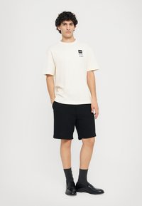 Cream short-sleeve T-shirt with a black logo patch, paired with black shorts, dark socks, and black shoes. Plain background.
