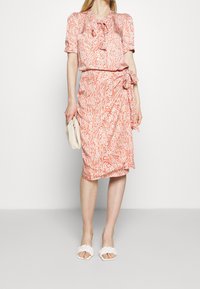 Šaty s květinovým vzorem a stahovacími pásky v jemných broskvových a bílých odstínech, doplněné bílými pletenými sandály na klínu. Texturovaná látka s nápadnými rukávy.