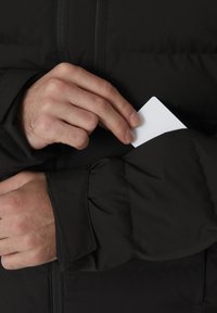 Schwarze Daunenjacke mit einem Reißverschluss vorne und Klettverschlüssen an den Ärmeln. Eine Hand steckt eine weiße Karte in eine Tasche am Ärmel. Glatte, matte Textur.