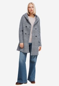 Dubbelzijdige houndstoothjas in zwart en wit, met een capuchon, gecombineerd met een lichte trui en wijde jeans.