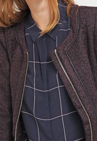 Vínová texturovaná bunda s černým lemováním a stříbrným zipem, nošená na tmavě modré košili s bílým mřížkovým vzorem a detaily na límci.