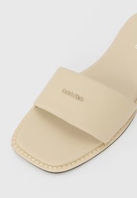 Beige Rutsch-Sandale mit einem breiten Riemen aus glattem Material, quadratischer Zehenform und dezenten Nähten am Rand sowie einem Logodetail.