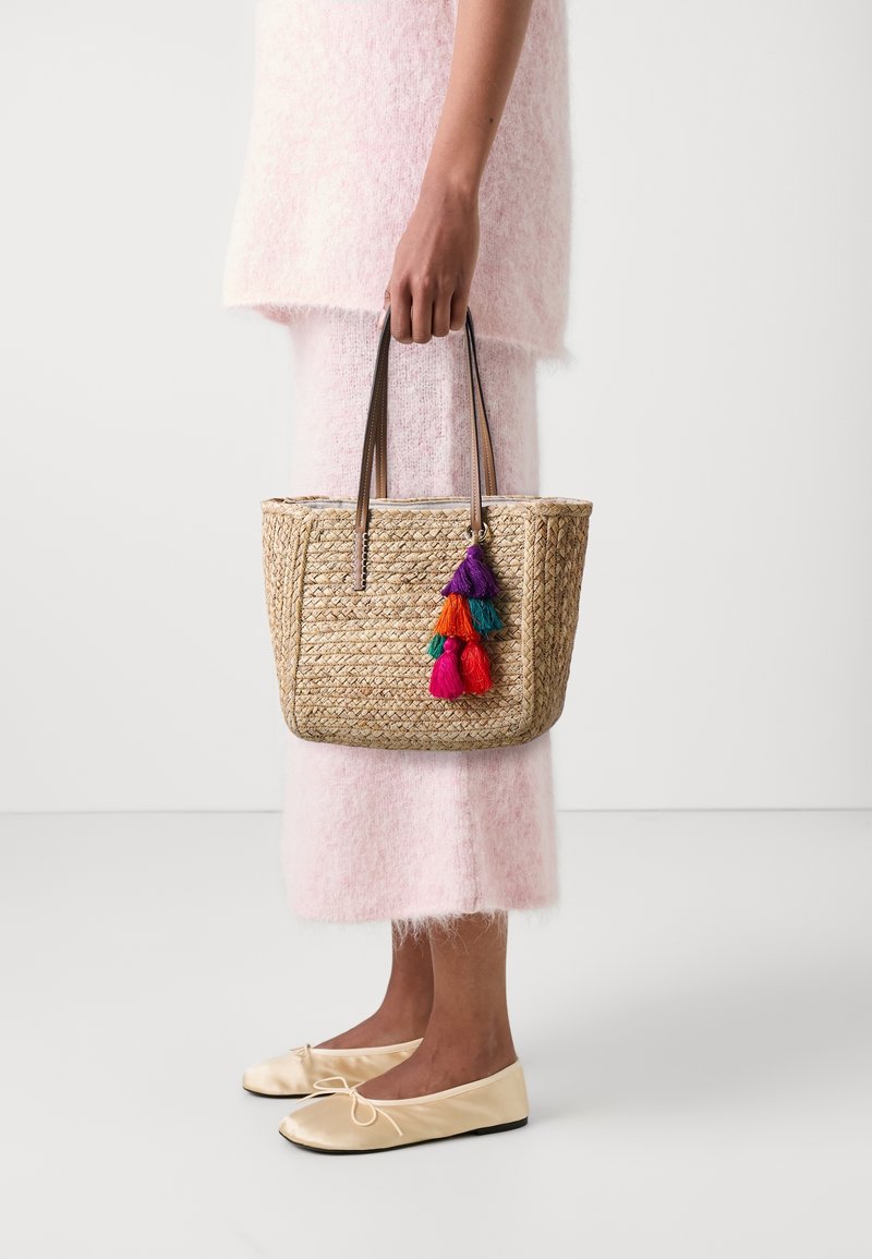 Personne en tenue rose pâle tenant un sac cabas en paille tressée avec des pompons colorés, portant des ballerines crème, debout sur un sol blanc.