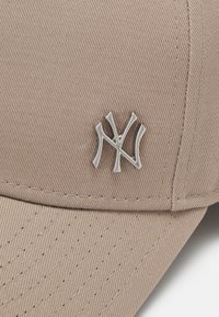 Gorra de béisbol beige con un acabado texturizado, que presenta un logotipo bordado en plata en la parte frontal. Los detalles de costura son visibles en la gorra.