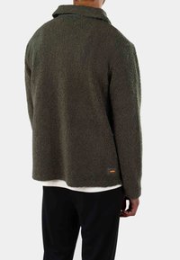 Giacca in pile verde con superficie texturizzata, colletto e una vestibilità leggermente oversize. Presenta un piccolo logo arancione sull'orlo.