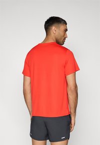 Κόκκινο αθλητικό μπλουζάκι με στρογγυλεμένο λαιμό, κοντά μανίκια και λεία υφή, συνδυασμένο με σκούρα σορτς που έχουν ένα λευκό διακριτικό στοιχείο.