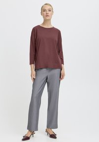 Bordeauxfarbene, übergroße Bluse mit drei Viertelärmeln, kombiniert mit grauen, maßgeschneiderten Hosen. Das Modell trägt bordeauxrote, spitze High Heels. Einfaches, klares Design.