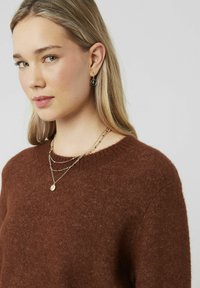 Un pull en tricot marron avec un col rond, des colliers en or superposés et une boucle d'oreille en or avec un charme noir.