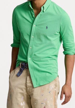 Man draagt een groen overhemd met lange mouwen, de mouwen opgerold, en beige broek met verfspatten en een gestreepte riem.