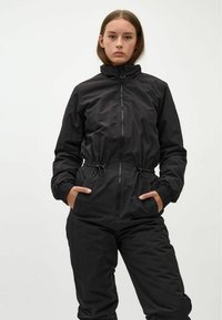 Schwarzer Jumpsuit mit Reißverschluss vorne, langen Ärmeln, elastischen Bündchen und verstellbarem Bund. Glatte Textur, minimalistisches Design mit seitlichen Taschen.