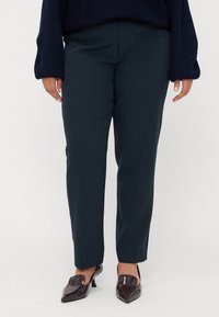 Navy blue tapered trousers made of smooth fabric, featuring a mid-rise waist and a clean, minimalist design. Worn with dark pointed shoes.