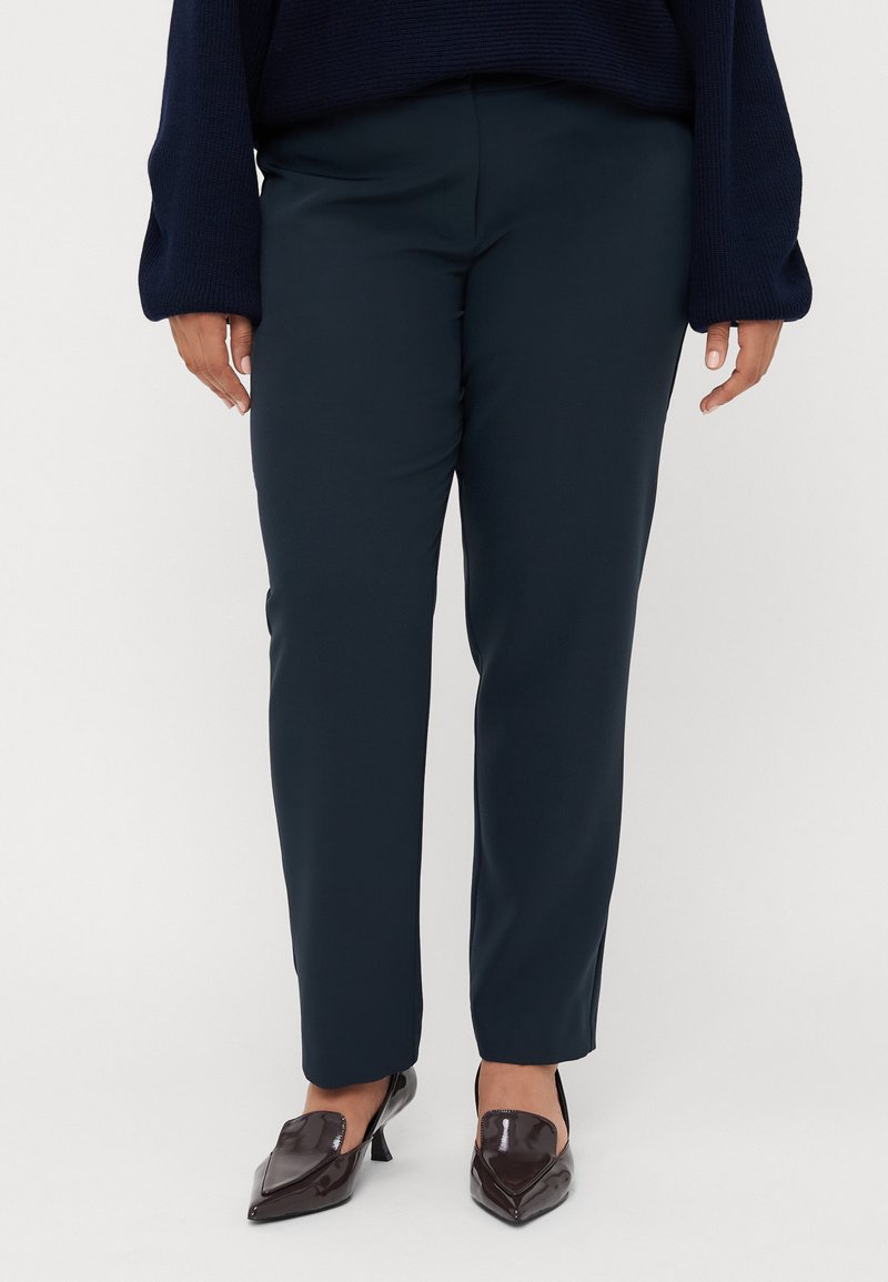 Navy blue tapered trousers made of smooth fabric, featuring a mid-rise waist and a clean, minimalist design. Worn with dark pointed shoes.
