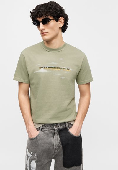 Ανοιχτό πράσινο βαμβακερό μπλουζάκι με στρογγυλή λαιμόκοψη και ξεθωριασμένο γραφικό σχέδιο. Φορεμένο με γκρι τζιν παντελόνι που έχει μαύρο μπαλώμα και σχέδιο στο πλάι.