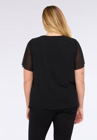 Donna bionda con capelli lunghi girata di spalle, indossa una maglietta nera a maniche corte con un sottile motivo a pois verticali e pantaloni neri.