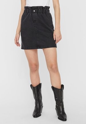 Mini-jupe en denim noir avec une taille froncée, fermeture à boutons sur le devant et poches sur les côtés, associée à des bottes en cuir noir hauteur genou.