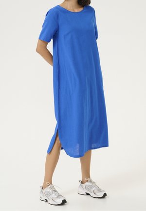 Femme portant une robe midi bleue ample avec des fentes latérales et des baskets blanches, debout les mains dans le dos.