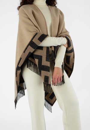Poncho in lana beige con motivo geometrico nero, dettagli in frange lungo l'orlo e design drappeggiato. Indossato sopra una maglia di colore chiaro.