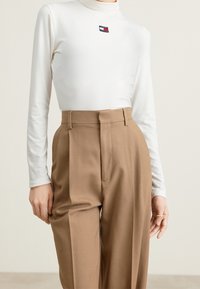 Woman wearing a white long-sleeve turtleneck with Tommy Jeans logo and high-waisted brown pleated trousers, ring on right hand.