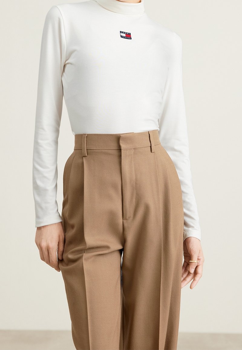 Woman wearing a white long-sleeve turtleneck with Tommy Jeans logo and high-waisted brown pleated trousers, ring on right hand.