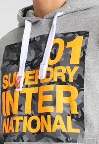 Sudadera gris con cordones blancos y un cuadrado de camuflaje negro y gris que presenta un gran texto amarillo que dice "01 Superdry International."