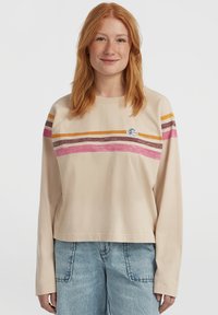 Young woman with red hair wearing beige long-sleeve shirt with orange, brown, and pink stripes and light blue jeans, standing against white background.