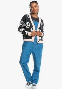 Black cardigan with white peace and abstract symbols, pink ribbed cuffs, and a button-up front, worn over denim overalls and canvas sneakers.