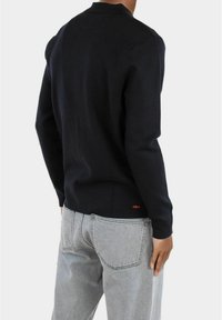 Maglioncino blu navy con scollo rotondo e maniche lunghe, abbinato a jeans in denim grigio chiaro. Presenta un piccolo logo rosso all'orlo.
