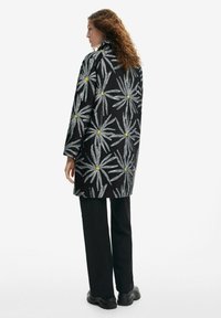 Black coat featuring a floral pattern in silver and yellow, made from textured fabric. Oversized design with long sleeves. Worn with black pants.