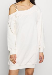Hvid off-shoulder sweatshirtklänning med långa ärmar, prydd med en dekorativ kedjesnodd och ribbad fåll. Tillverkad av mjukt bomullsmaterial.