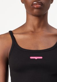 Canottiera nera a costine con logo "tommyjeans" rosa sul petto. Le spalline sono sottili e il materiale ha una consistenza morbida.