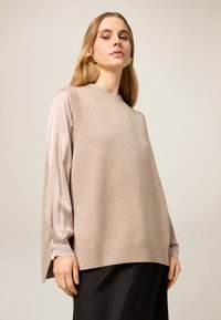 Beiger ärmelloser Strickpullover mit geripptem Ausschnitt und Saum, getragen über einer hellrosa Satinbluse mit geknöpften Manschetten, kombiniert mit schwarzen Hosen.