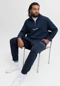 Mörkblå zip-up sweatshirt med vitt logo och matchande joggers, tillverkade av mjukt tyg. Vita sneakers med svarta detaljerade strumpor på en stol.