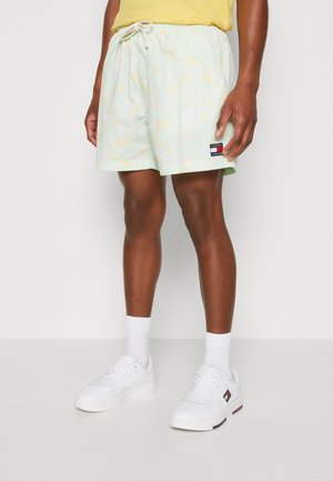 Hombre vistiendo pantalones cortos de camuflaje en tonos pastel claros con el logo de Tommy Jeans, calcetines blancos y zapatillas blancas con un emblema de la bandera Tommy.