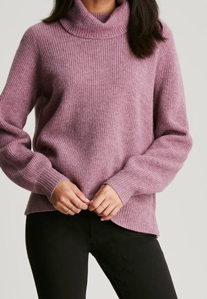 Femme portant un pull à col roulé côtelé mauve à manches longues avec un pantalon noir, les mains croisées au niveau de la taille.