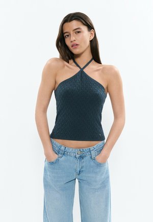 Femme aux longs cheveux bruns portant un haut dos nu bleu foncé texturé et un jean taille haute bleu clair, les mains dans les poches.