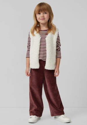 Un gilet blanc en peluche sur une chemise à manches longues à rayures marron et blanche, associé à un pantalon large en velours côtelé bordeaux et des baskets blanches.