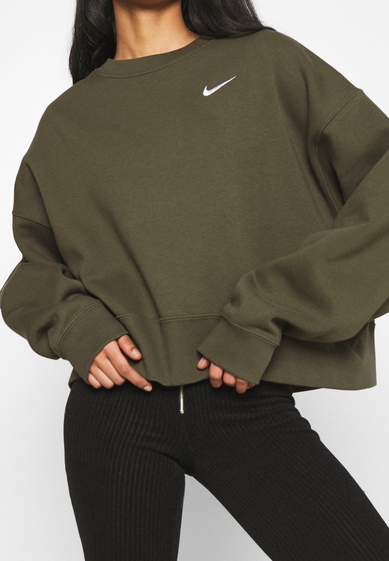 Oliivinvihreä lyhyt huppari pitkillä hihoilla ja pyöreällä kaula-aukolla, jossa on pieni valkoinen Nike-logo rinnassa. Mustat ribbitun näköiset housut.