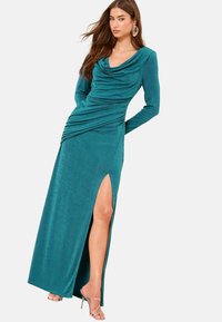 Dlhé šaty modrozelené farby s raglánovým dlhým rukávom, s raglánovým golierom a bočným rozparkom. Vyrobené zo štruktúrovanej látky; majú nariasenie v páse a minimálne kovové detaily.