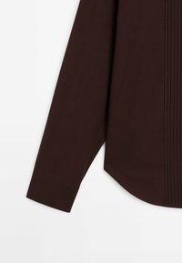 Chemise à manches longues marron foncé avec un design structuré, comprenant des plis verticaux, un poignet à boutons et un ourlet légèrement courbé.