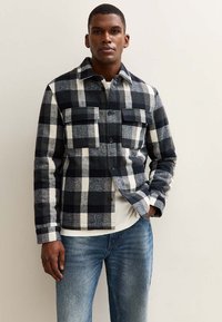 Mann trägt eine schwarz-weiß-graue karierte Button-Up-Jacke mit zwei Brusttaschen und blauen Jeans, steht gegen eine helle Wand.