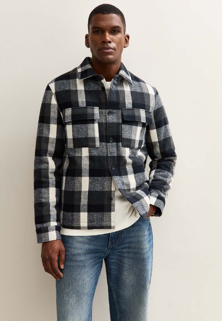 Mann trägt eine schwarz-weiß-graue karierte Button-Up-Jacke mit zwei Brusttaschen und blauen Jeans, steht gegen eine helle Wand.