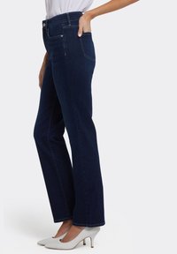 Donkerblauwe denim jeans met rechte pijpen en een hoge taille, voorzien van zakken en subtiele stikdetails, gecombineerd met lichtgrijze hoge hakken.