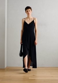 Robe noire sans manches avec un décolleté drapé, un ourlet asymétrique et des bretelles en chaîne argentée. Portée avec des chaussures noires et un petit sac.