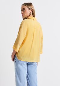 Camicia gialla, leggera, con una consistenza morbida e trasparente, dotata di colletto e maniche arrotolate, abbinata a jeans azzurri chiari e comodi.