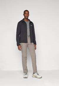 Giacca nera con zip e una t-shirt grigia sotto, abbinata a pantaloni cargo beige e scarpe da ginnastica bianche con dettagli neri.