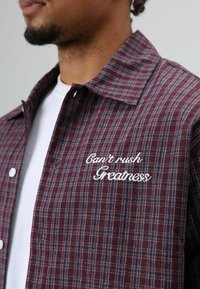 Chemise à carreaux rouge et bleue, avec une phrase brodée en blanc "Can't rush Greatness" sur le côté gauche de la poitrine. Col boutonné, en coton.