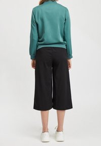 Femme portant un haut à manches longues bleu sarcelle, un pantalon large noir raccourci et des baskets blanches, debout de dos sur un fond uni.