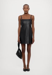 Rochie din piele neagră cu decolteu pătrat și bretele subțiri, având o siluetă ajustată. Modelul ține o geantă neagră asortată.