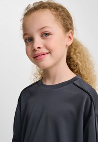 Grå träningströja med en strukturerad tyg, rund halsringning och raglanärmar, bärs av ett barn med lockigt hår. Mjuk, avslappnad design.